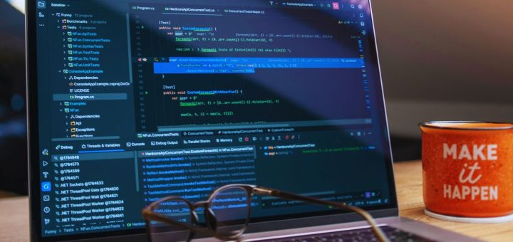 Laptop displaying a dark code editor with multiple panels and blue syntax highlighting; glasses rest on the keyboard and an orange mug reading 'MAKE it HAPPEN' sits to the right on the desk.