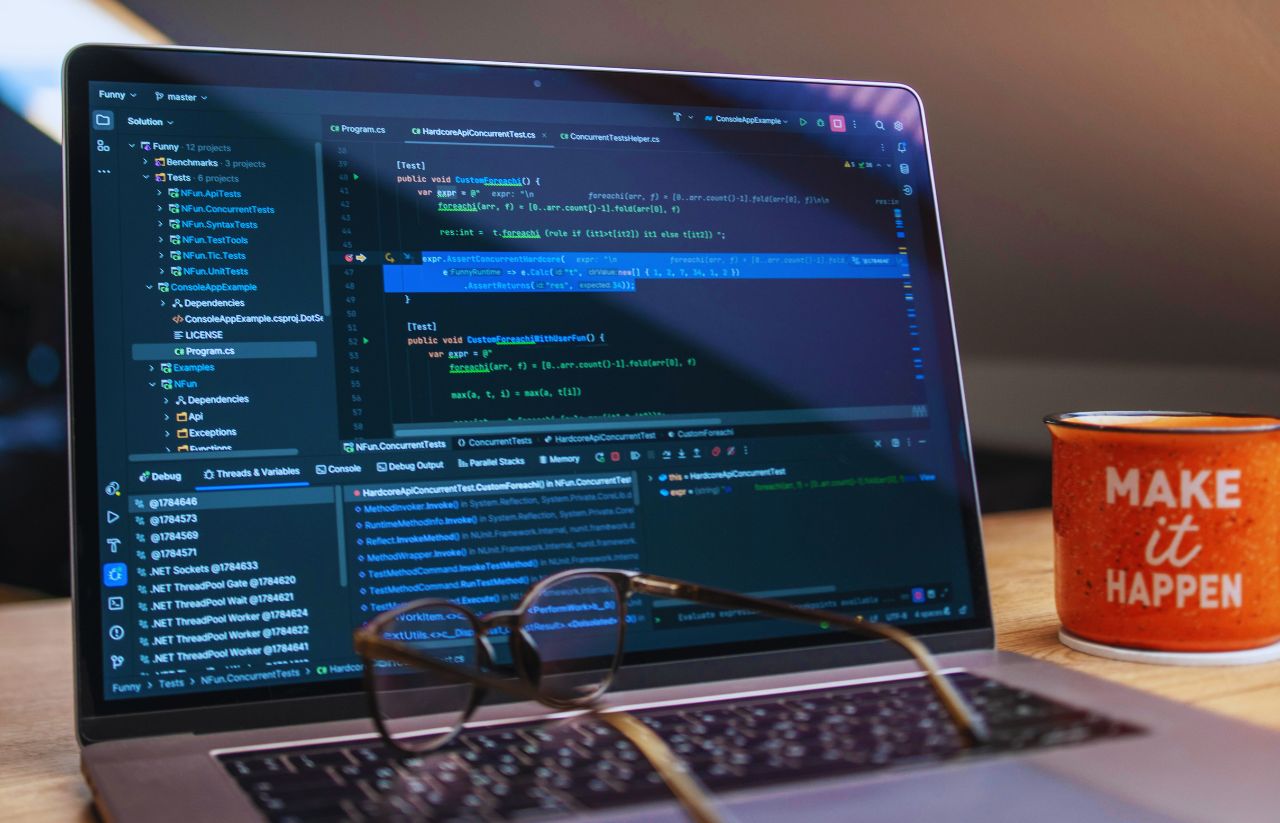 Laptop displaying a dark code editor with multiple panels and blue syntax highlighting; glasses rest on the keyboard and an orange mug reading 'MAKE it HAPPEN' sits to the right on the desk.