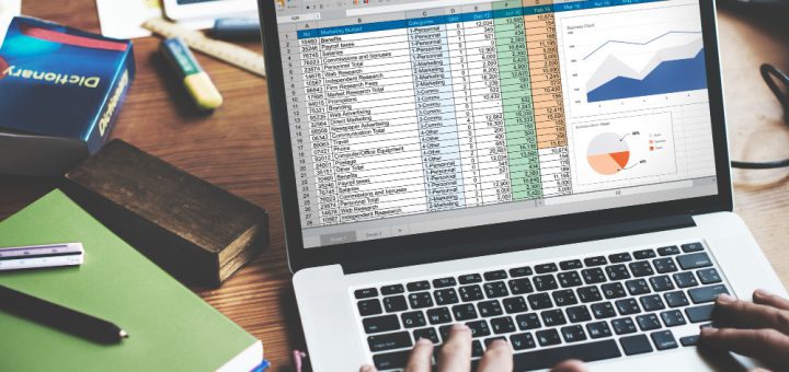Open laptop showing a detailed spreadsheet with tables and two charts on a desk, surrounded by notebooks and office supplies.