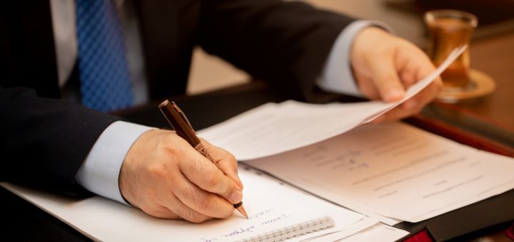 Businessperson in a suit signs documents at a desk with a brown pen, papers spread out and a cup on the side