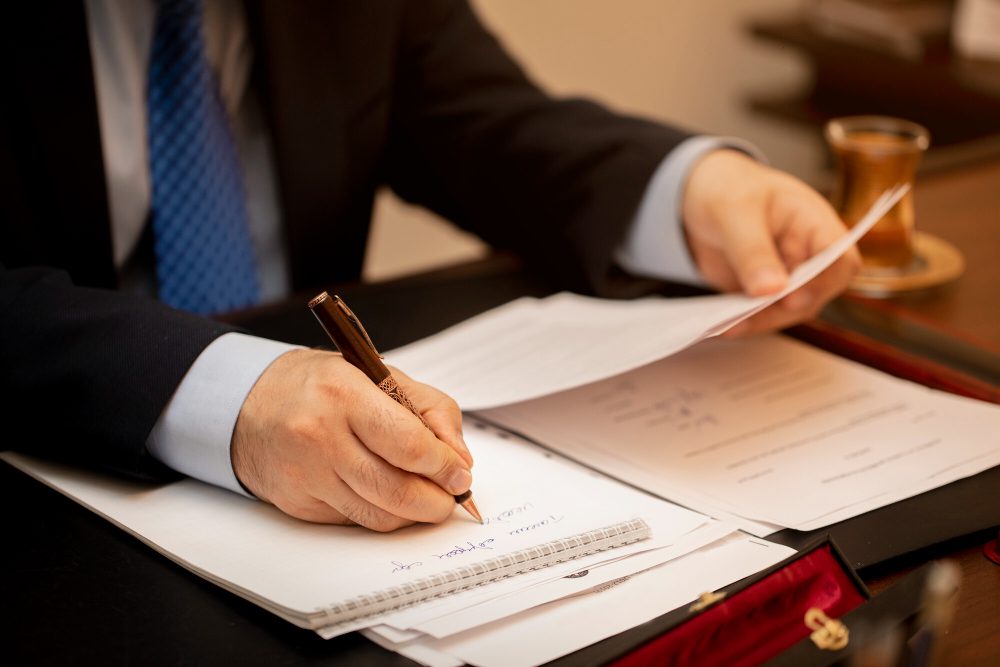 Businessperson in a suit signs documents at a desk with a brown pen, papers spread out and a cup on the side