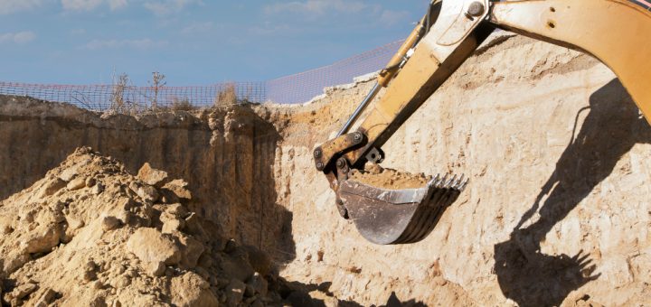 Excavator bucket digging into a sandy trench at a construction site, with piled soil and blue sky beyond.