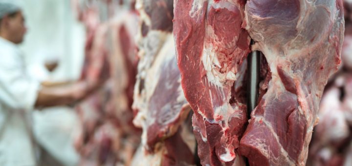 Hanging slabs of raw red meat in a butcher shop with a worker in the background.