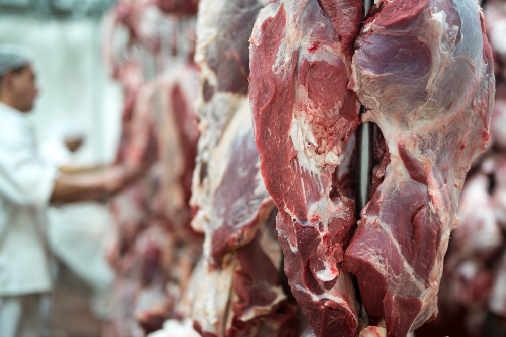Hanging slabs of raw red meat in a butcher shop with a worker in the background.