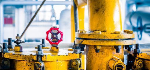 Close-up of a yellow industrial valve assembly with a red handwheel in a factory setting, showing bolts and pipes nearby.