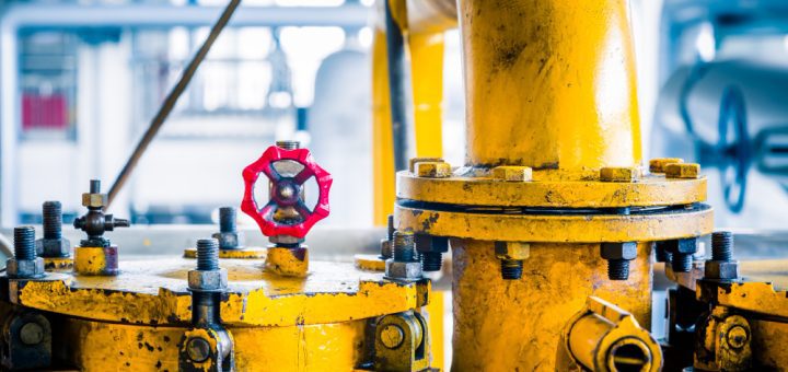 Close-up of a yellow industrial valve assembly with a red handwheel in a factory setting, showing bolts and pipes nearby.