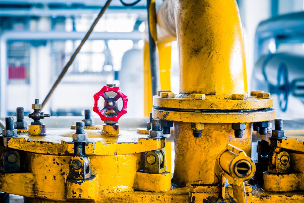 Close-up of a yellow industrial valve assembly with a red handwheel in a factory setting, showing bolts and pipes nearby.