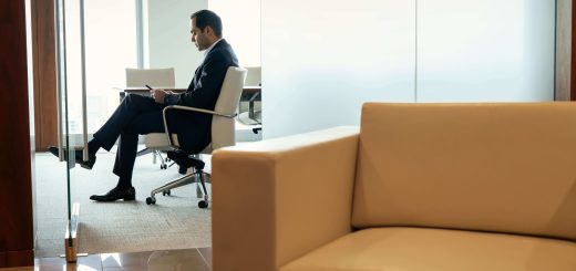 Businessman in a dark suit sits in a white office chair, reading documents at a desk beside a glass partition, with a beige sofa in the foreground.