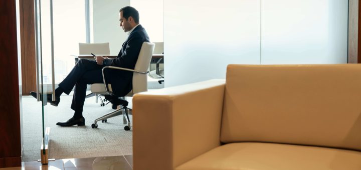 Businessman in a dark suit sits in a white office chair, reading documents at a desk beside a glass partition, with a beige sofa in the foreground.