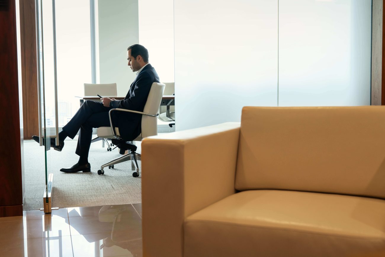 Businessman in a dark suit sits in a white office chair, reading documents at a desk beside a glass partition, with a beige sofa in the foreground.
