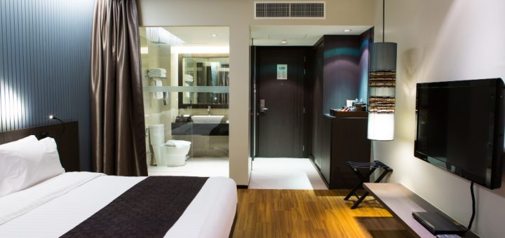 Hotel room with king bed, wall-mounted headboard, and dark brown runner; glass-walled bathroom to the left, TV on wall to the right, wooden floor and modern lighting.