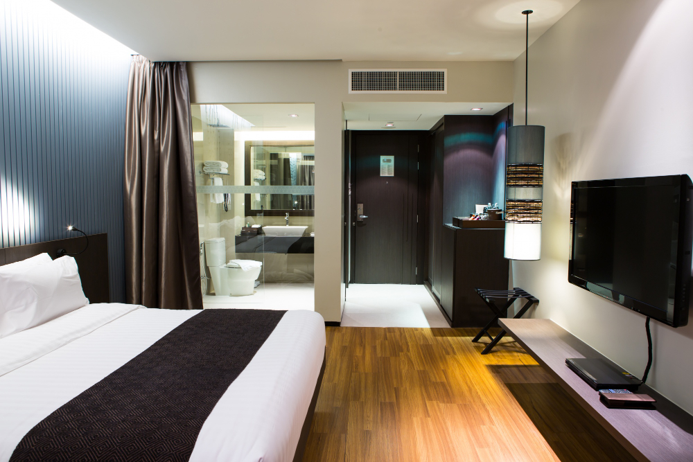 Hotel room with king bed, wall-mounted headboard, and dark brown runner; glass-walled bathroom to the left, TV on wall to the right, wooden floor and modern lighting.