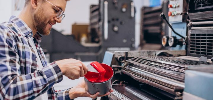 Print shop worker in a plaid shirt and safety glasses pours red ink from a can into a container beside a large printing press.