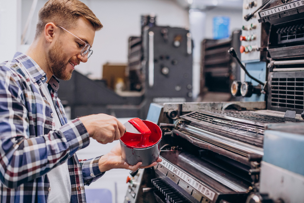 Print shop worker in a plaid shirt and safety glasses pours red ink from a can into a container beside a large printing press.