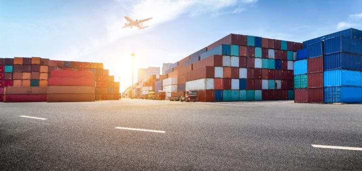 Shipping yard with stacked colorful cargo containers and a plane taking off in a bright blue sky