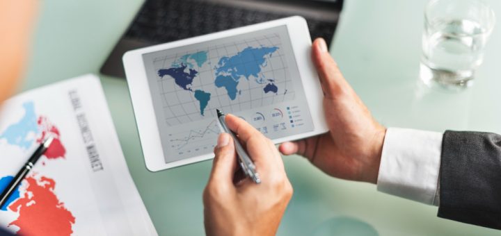 Person in a suit uses a tablet displaying a world map and charts on a desk with documents nearby and a glass of water nearby.