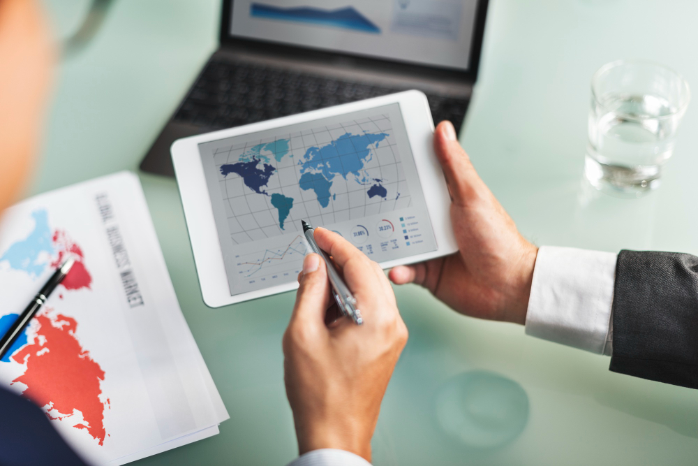 Person in a suit uses a tablet displaying a world map and charts on a desk with documents nearby and a glass of water nearby.