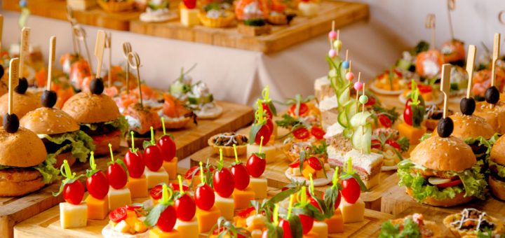 Assorted bite-size appetizers on wooden boards at a catered event, featuring sliders, cheese cubes with cherry tomatoes, and colorful skewers.