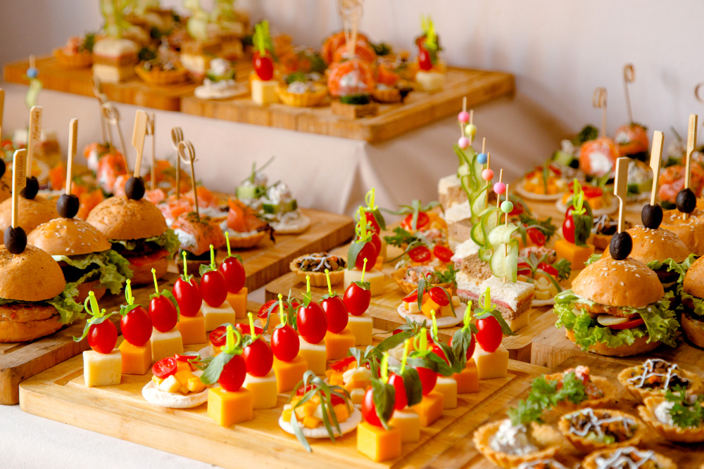 Assorted bite-size appetizers on wooden boards at a catered event, featuring sliders, cheese cubes with cherry tomatoes, and colorful skewers.