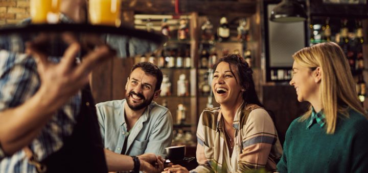 Three friends seated at a bar, smiling and chatting as a server pours drinks in the foreground.
