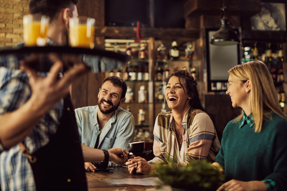773073 - AS Harmony | Negocios10.Com Three friends seated at a bar, smiling and chatting as a server pours drinks in the foreground.