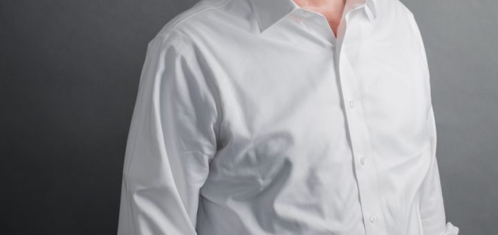 Professional head-and-shoulders portrait of a middle-aged man in a white shirt and dark trousers, looking at the camera against a gray backdrop.