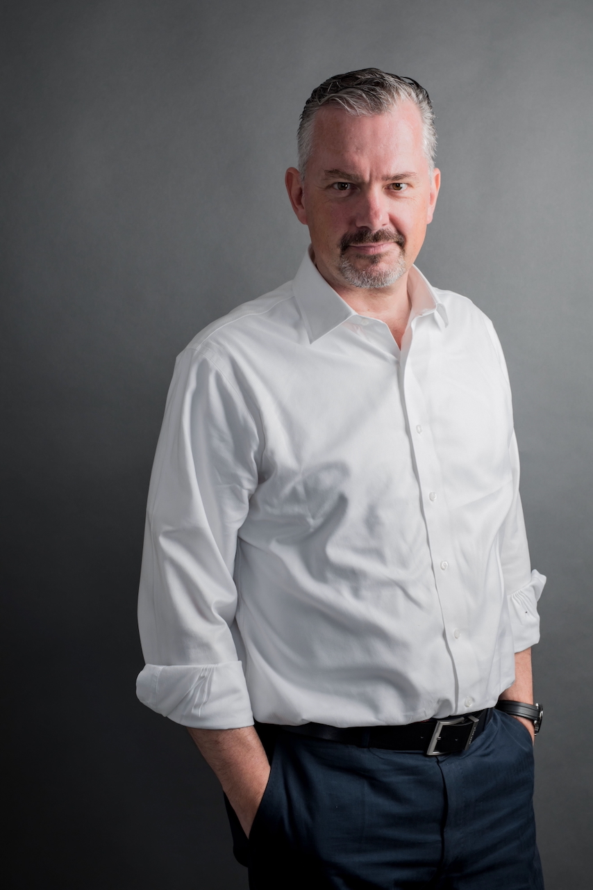 Professional head-and-shoulders portrait of a middle-aged man in a white shirt and dark trousers, looking at the camera against a gray backdrop.