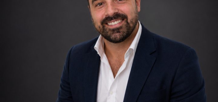 Portrait of a smiling man in a navy blazer and white shirt against a dark studio backdrop.