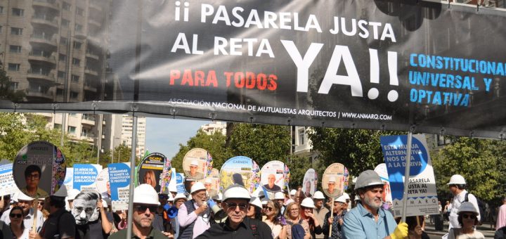 Crowd of protesters wearing hard hats march under a large banner that reads '¡ PASARELA JUSTA AL RETA YA!' with round placards above them in a city street constraints over.