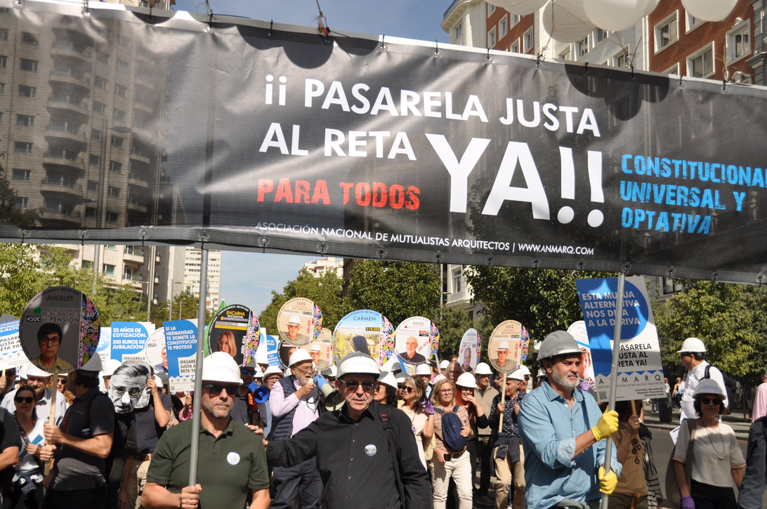 Mutualistas - IP | Negocios10.Com Crowd of protesters wearing hard hats march under a large banner that reads '¡ PASARELA JUSTA AL RETA YA!' with round placards above them in a city street constraints over.