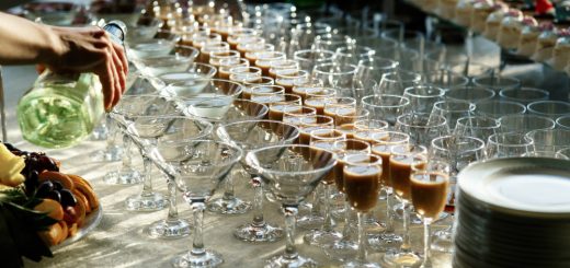 Person pouring champagne into a row of fluted glasses on a catered buffet table with fruit and desserts nearby.