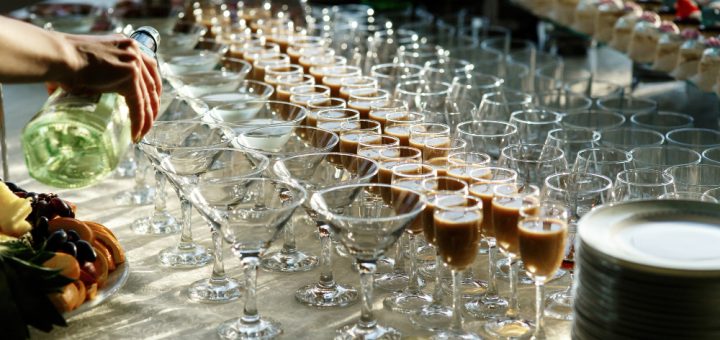 Person pouring champagne into a row of fluted glasses on a catered buffet table with fruit and desserts nearby.