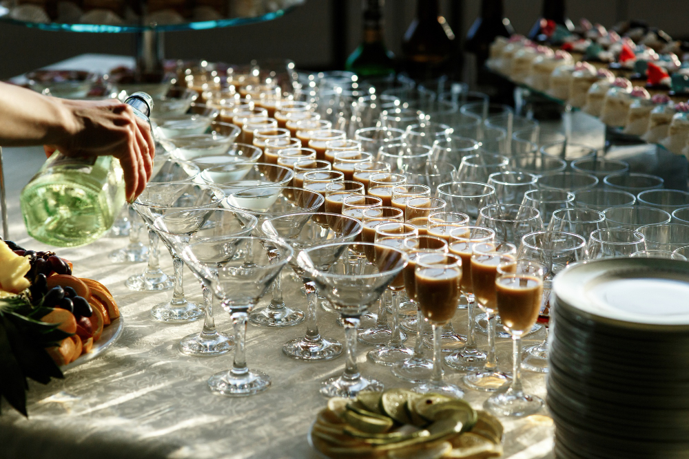 773131 - Santa Jarana | Negocios10.Com Person pouring champagne into a row of fluted glasses on a catered buffet table with fruit and desserts nearby.