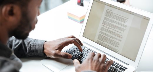 Close-up of a person typing on a silver laptop at a white desk, screen displaying a dense document