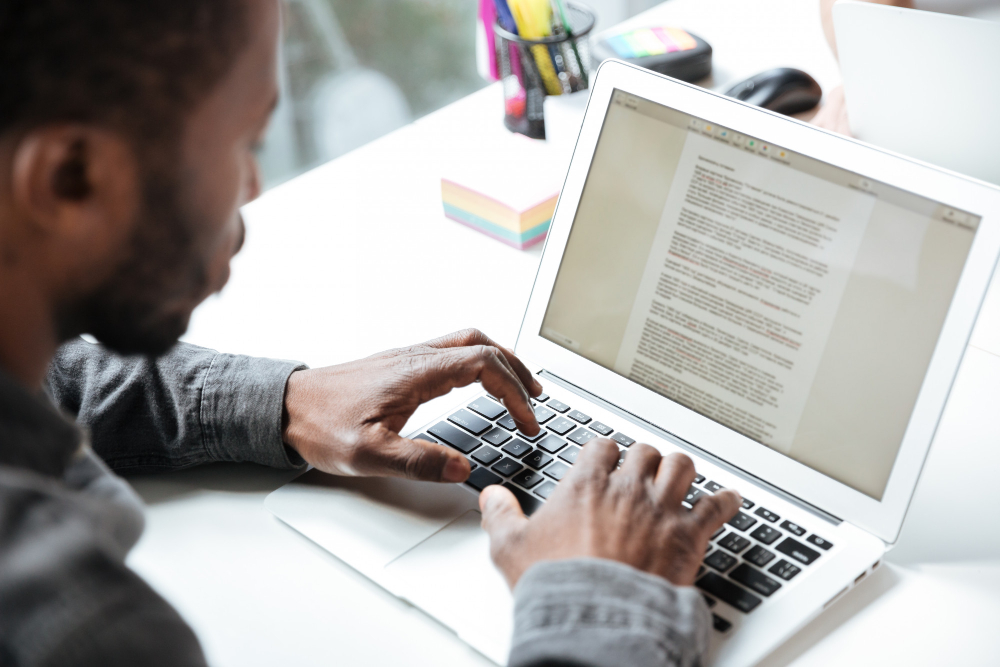 773757 - Si Quieres te lo Escribo | Negocios10.Com Close-up of a person typing on a silver laptop at a white desk, screen displaying a dense document