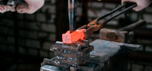 Blacksmith forging a bright orange-hot metal bar in a vise, wearing gloves.