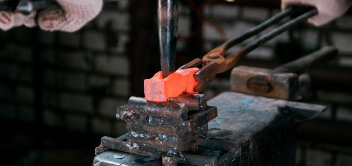 Blacksmith forging a bright orange-hot metal bar in a vise, wearing gloves.