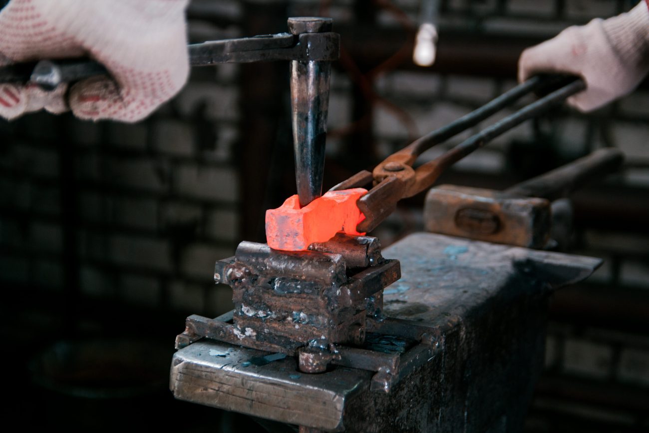 Blacksmith forging a bright orange-hot metal bar in a vise, wearing gloves.