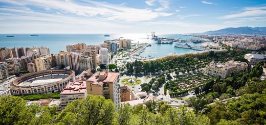 Panoramic view of a coastal city with a harbor, high-rise buildings, and a green park in the foreground under a clear blue sky to the horizon.