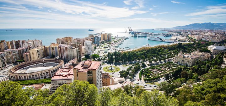 Panoramic view of a coastal city with a harbor, high-rise buildings, and a green park in the foreground under a clear blue sky to the horizon.