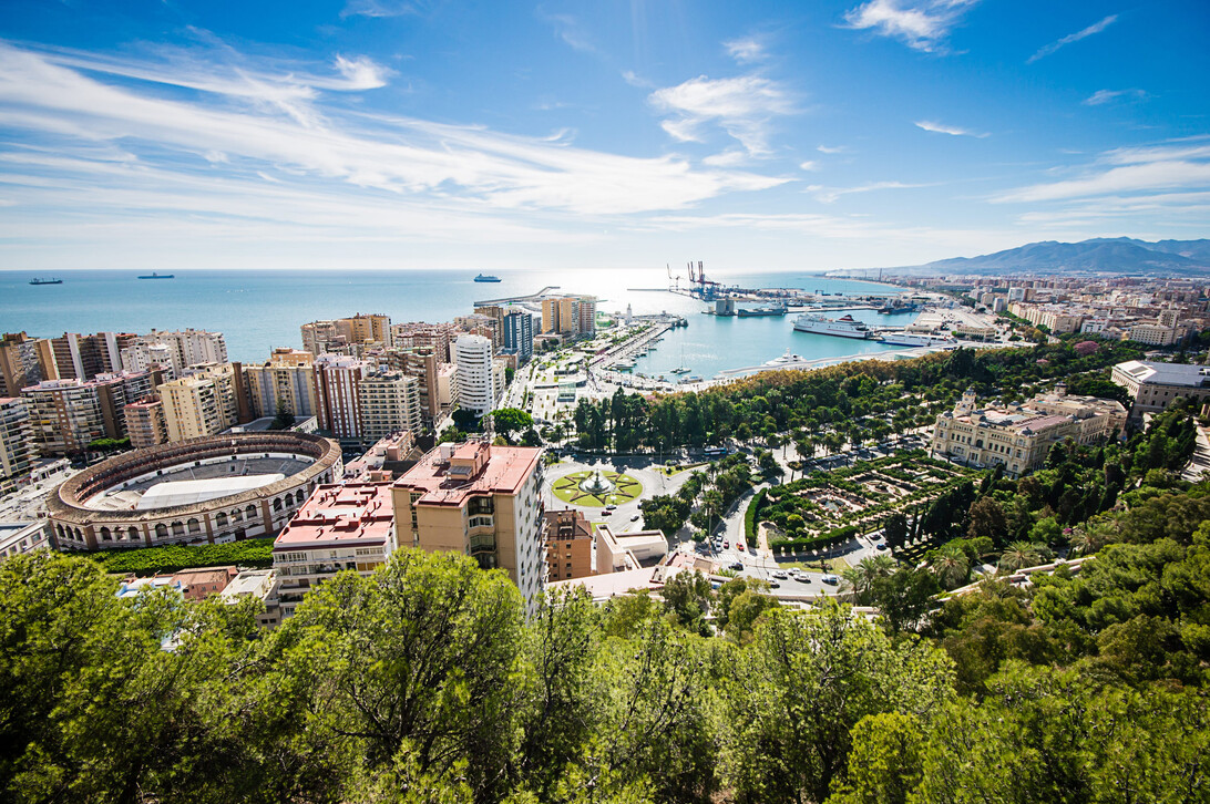 Despachos malaga - IP | Negocios10.Com Panoramic view of a coastal city with a harbor, high-rise buildings, and a green park in the foreground under a clear blue sky to the horizon.