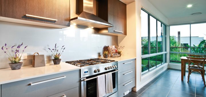 Modern kitchen with stainless steel range and range hood, gray lower cabinets, wood upper cabinets, white countertops, and large windows overlooking a patio and garden.