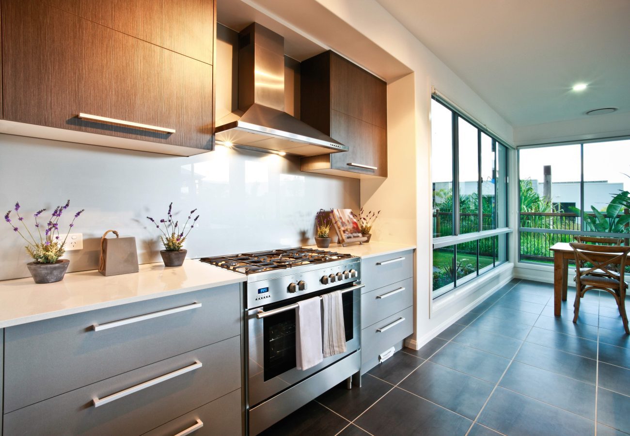 777554 - Carpintería Molas | Negocios10.Com Modern kitchen with stainless steel range and range hood, gray lower cabinets, wood upper cabinets, white countertops, and large windows overlooking a patio and garden.