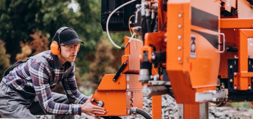 Worker in a plaid shirt and orange ear protection operates an orange industrial log-cutting machine outdoors, adjusting the control.