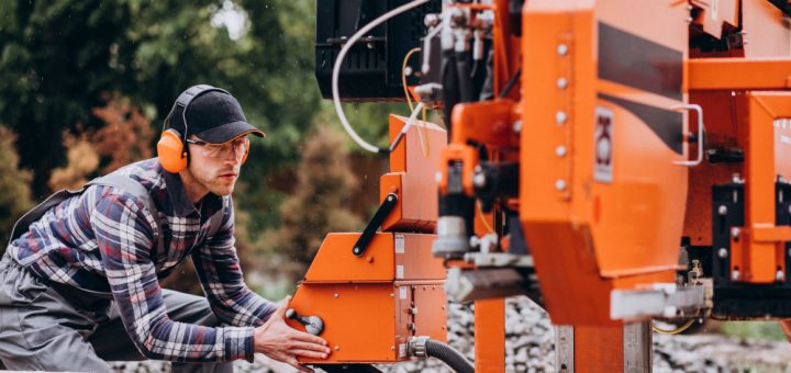 Worker in a plaid shirt and orange ear protection operates an orange industrial log-cutting machine outdoors, adjusting the control.