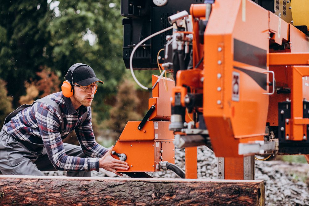 Worker in a plaid shirt and orange ear protection operates an orange industrial log-cutting machine outdoors, adjusting the control.