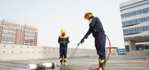 Two construction workers in yellow helmets and protective gear on a rooftop, one operating a power tool while the other watches over a rolled material nearby.
