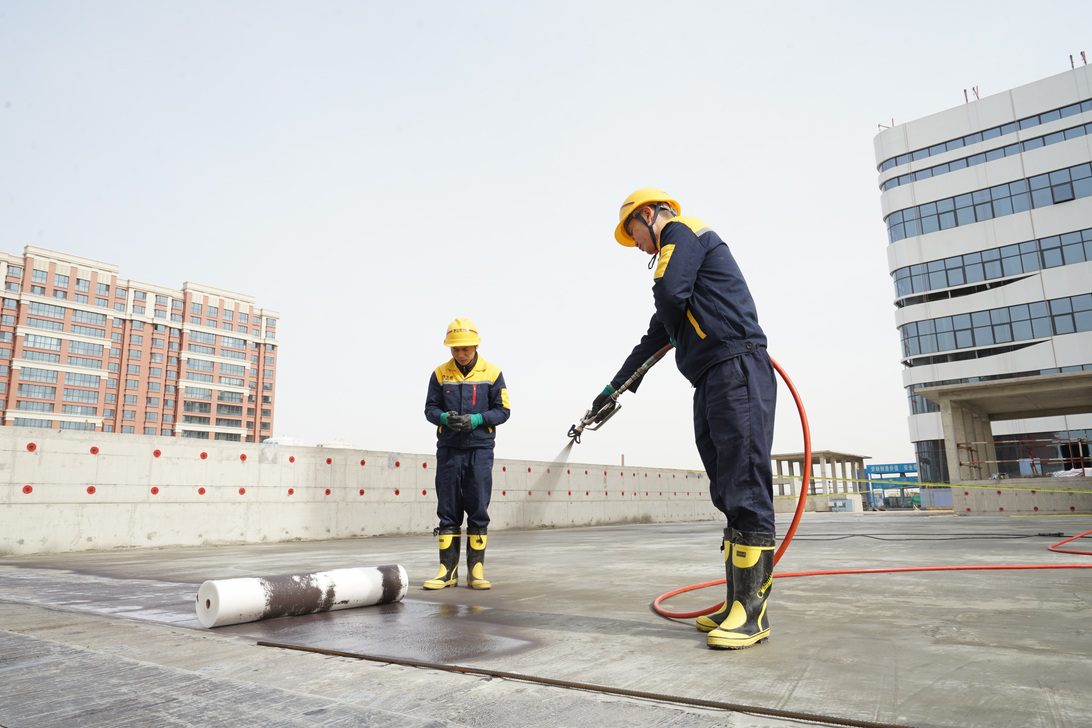 777977 - CINAT | Negocios10.Com Two construction workers in yellow helmets and protective gear on a rooftop, one operating a power tool while the other watches over a rolled material nearby.