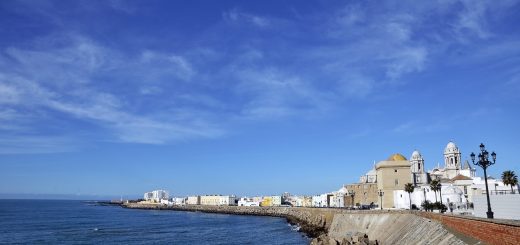 Coastal town along a rocky seawall under a clear blue sky, with white and beige buildings and domed church façades near the water