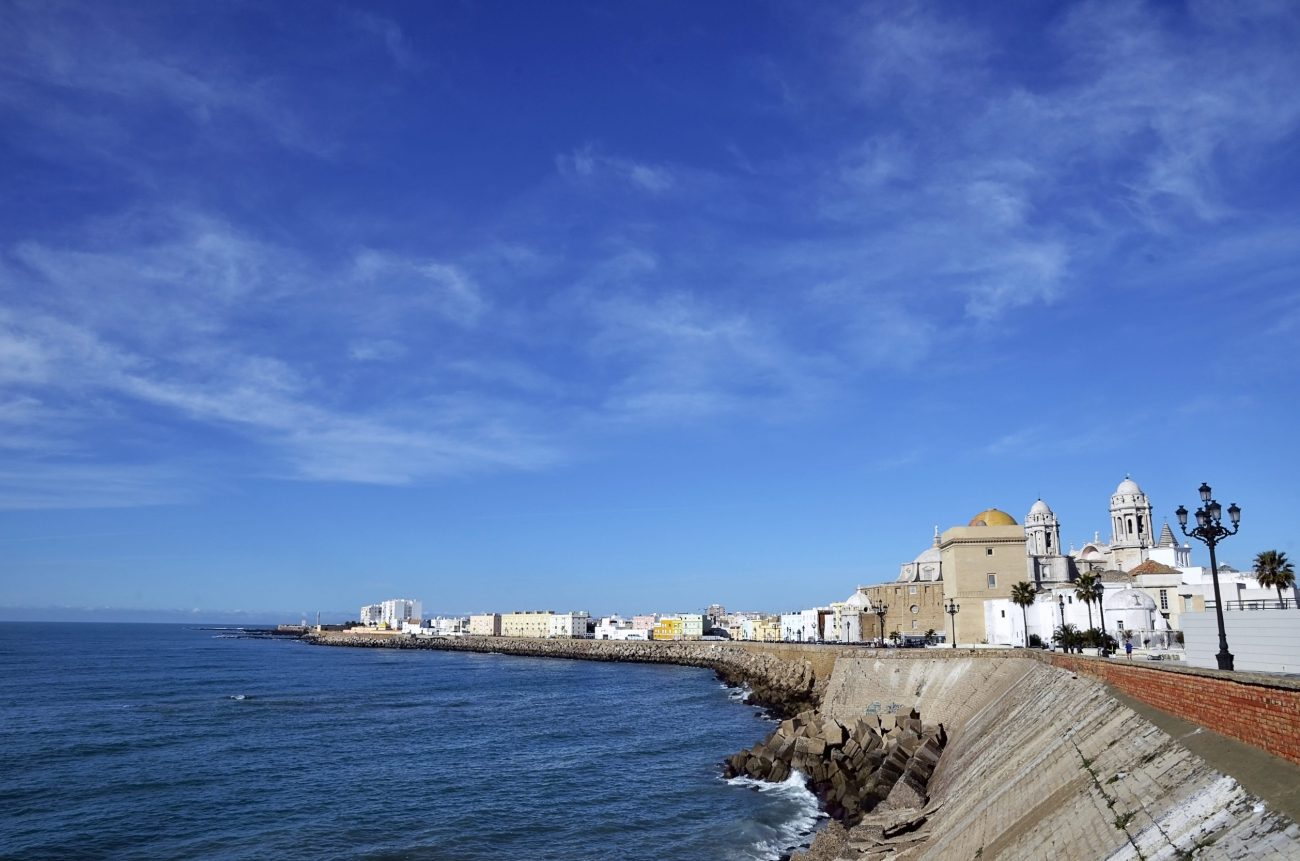 CADIZ SPAIN | Negocios10.Com Coastal town along a rocky seawall under a clear blue sky, with white and beige buildings and domed church façades near the water